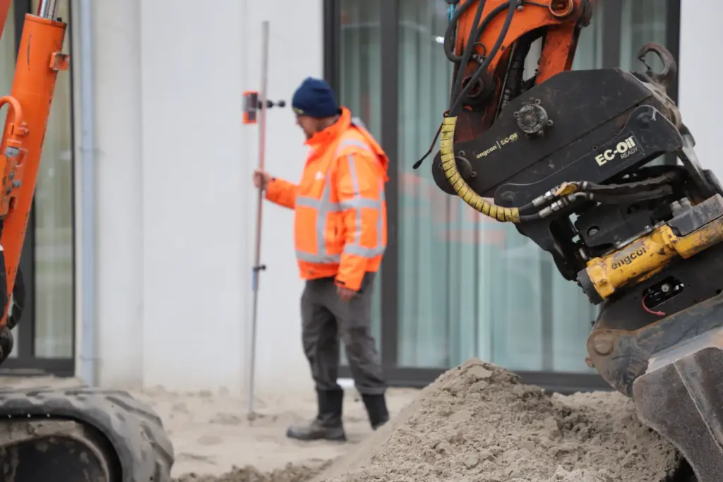 Vakman met meetapparatuur naast een graafmachine